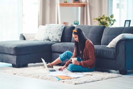 Young woman sits on the floor in a living room with a laptop and a cup of coffee going over documents