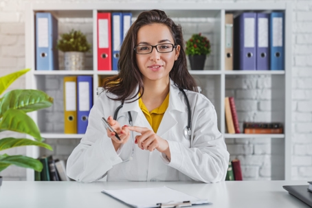 A woman doctor in a white coat is smiling and holding a pen.