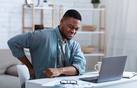 Young man sits at a home office desk clutching his lower back in pain