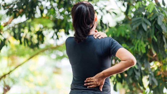 Young woman stands outdoors and holds her neck and lower back in pain