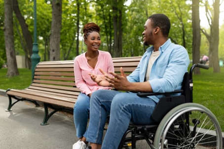 Young man in a wheelchair sits next to a woman on a park bench talking and smiling in the park