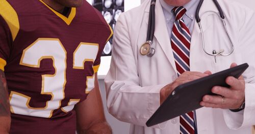 A doctor in a white coat is looking at a tablet, likely medical images, with a football player in a maroon uniform.