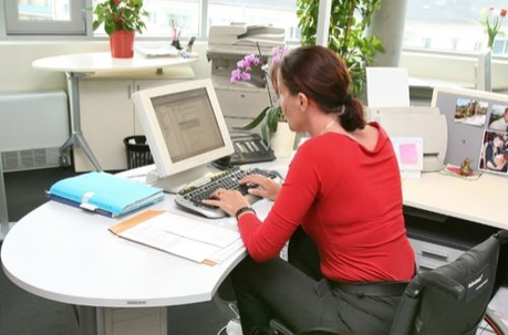A woman in a red shirt and wheelchair is working at a computer in an office.