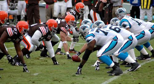 Football players in brown and blue uniforms are lined up on a field, ready to play. A football is in the middle.